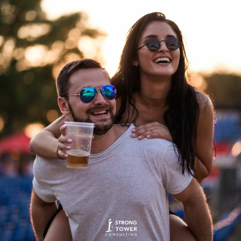 Woman riding on the a man's back with a drink in her hand at an outdoor event. 