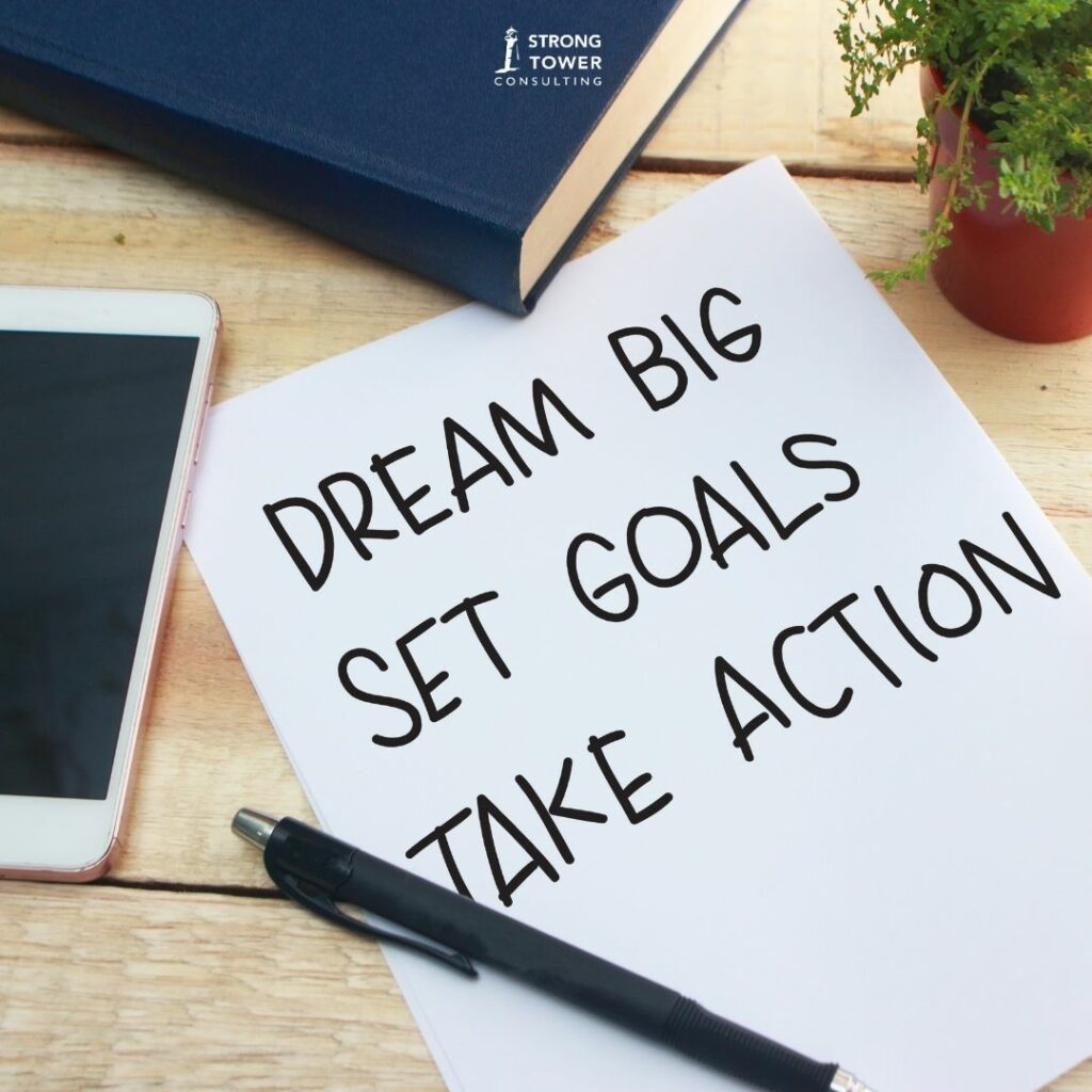 Desktop with plant, blue book, iPhone, and a paper with pen. The paper reads, "Dream big, set goals, take action."