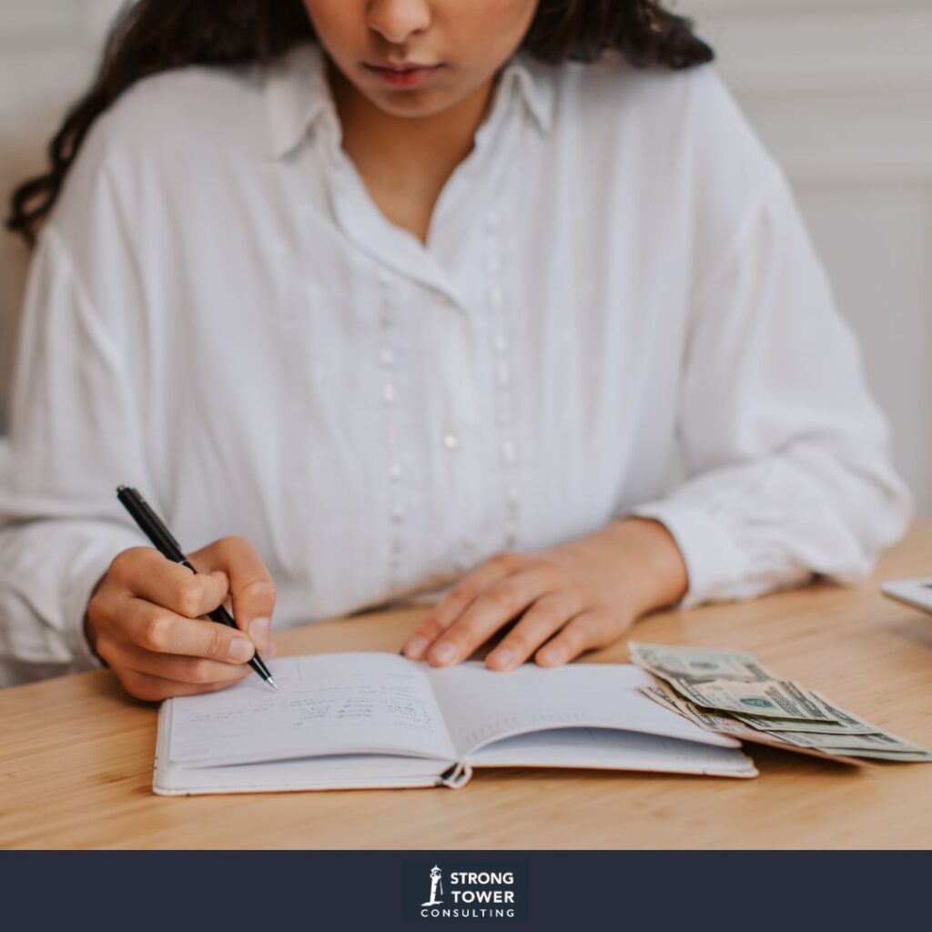 Woman in a white button-up filling out her budget on pen and paper with her cash close by